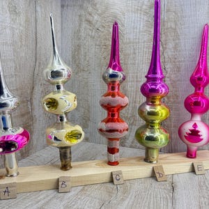 Può includere: Cinque puntali per albero di Natale in vetro vintage disposti su un supporto in legno etichettato da A a E. I puntali presentano forme varie, tra cui estremità appuntite e sezioni bulbose in argento, crema, rosso, verde, giallo e rosa acceso.