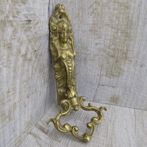 May include: A gold-toned metal wall hook with an ornate design featuring a woman's head and body. The hook is shaped like a curved bracket with a decorative scrollwork design.