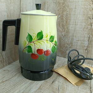 May include: Vintage electric coffee percolator with a black base and handle. The cream-colored body features a hand-painted design of red cherries and green leaves. A black power cord and plug are included.