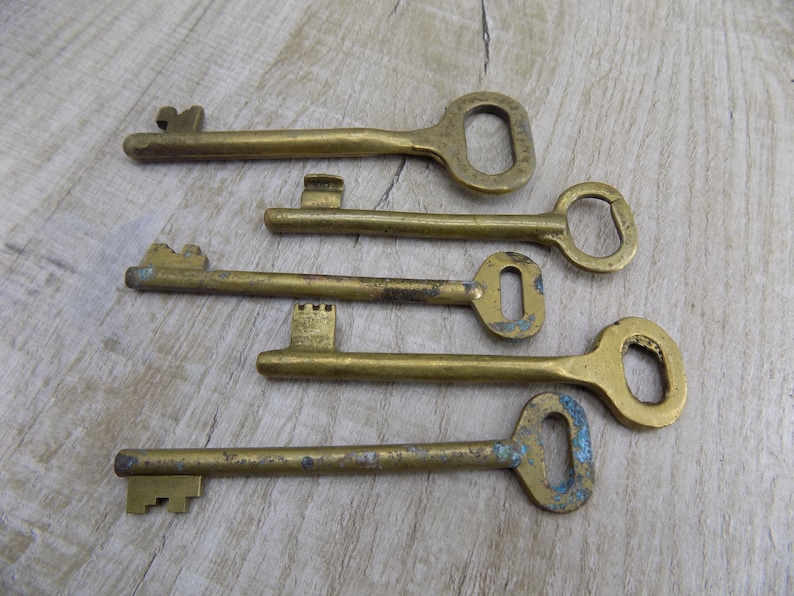 May include: Five antique brass keys with different shapes and sizes. The keys have a worn, aged look and are arranged on a light wood surface.