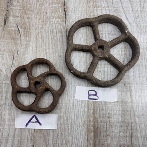 Peut inclure: Deux volants de vanne antiques en métal rouillé, probablement issus de machines industrielles. L'un est plus petit avec un design à six rayons, l'autre est plus grand avec un design à cinq rayons. Les deux sont étiquetés "A" et "B".