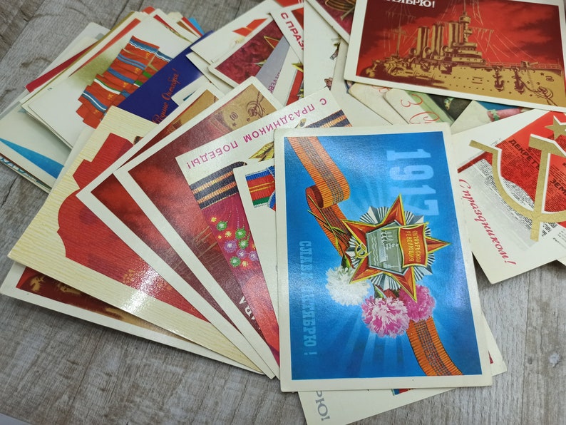 May include: A collection of vintage postcards with various designs and text. The cards feature red, blue, and white colors, with some showcasing military themes and historical imagery. The text includes dates and celebratory phrases.
