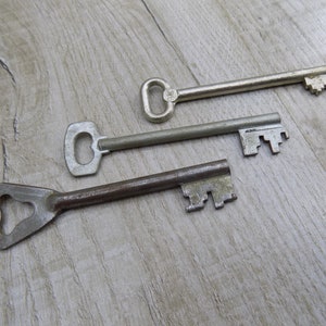 May include: Three antique keys on a light wood surface. The keys are made of metal and have different shapes and sizes. The key on the top is silver and has a simple design. The middle key is also silver and has a more intricate design. The bottom key is rusty and has a unique shape.