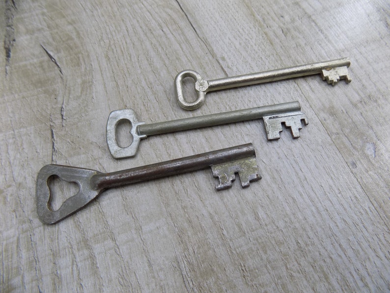 May include: Three antique keys on a light wood surface. The keys are made of metal and have different shapes and sizes. The key on the top is silver and has a simple design. The middle key is also silver and has a more intricate design. The bottom key is rusty and has a unique shape.