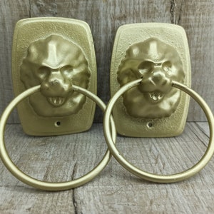May include: Two gold lion head towel rings with a smooth, shiny finish. Each ring has a lion head design with a detailed face and a circular ring.