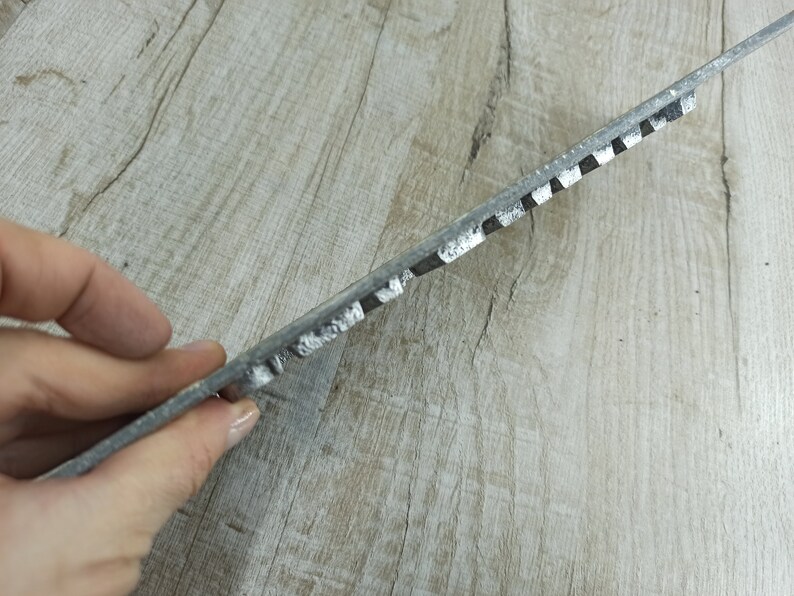 Puede incluir: Primer plano de una rejilla de metal gris con una serie de crestas elevadas. La rejilla se sostiene en una mano, y el fondo es una superficie de madera clara.