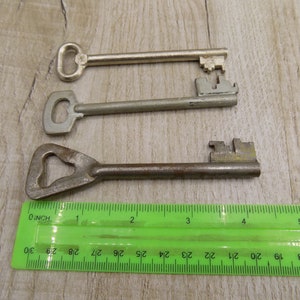 May include: Three keys of different sizes and styles, laid out on a wooden surface with a green ruler for scale. The keys are made of metal and have different shaped heads.