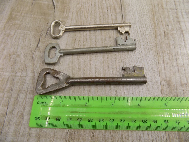 May include: Three keys of different sizes and styles, laid out on a wooden surface with a green ruler for scale. The keys are made of metal and have different shaped heads.