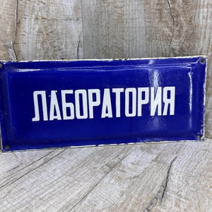 May include: A rectangular blue sign with the white word "ЛАБОРАТОРИЯ" (Laboratory) in capital letters. The sign has a white border and is mounted on a wooden surface.