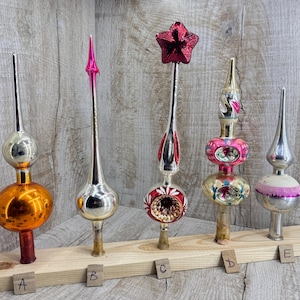 Può includere: Cinque puntali per albero di Natale in vetro vintage etichettati da A a E, disposti su una tavola di legno. Presentano varie forme, tra cui sfere, punte allungate e una stella, con finiture metalliche nei colori arancione, argento, rosso e rosa.