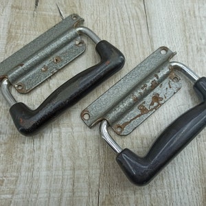 Pode incluir: Duas alças de mala vintage de metal com pegas de plástico preto. As alças são prateadas com um acabamento ligeiramente enferrujado.