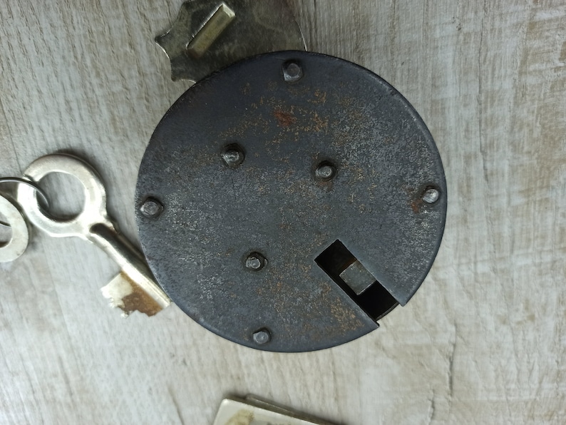 May include: A black metal lock with a keyhole and several rivets. The lock is on a light wood surface.