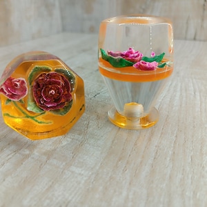 May include: Two clear glass decorative objects with floral designs. The larger object is shaped like a vase and the smaller object is shaped like a rounded dome. Both objects have a yellow base and feature pink and green flowers.