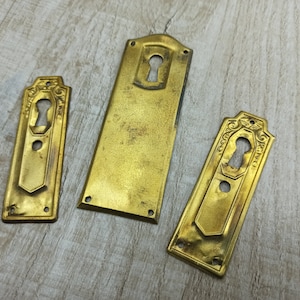 May include: Three antique brass keyhole escutcheons with ornate designs. The escutcheons are in various sizes and have a worn, aged finish.