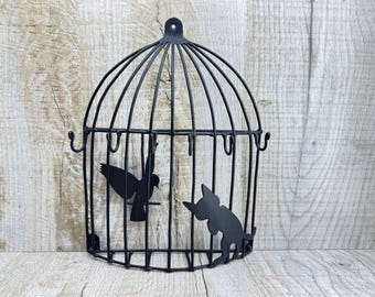 May include: Black metal decorative birdcage with a bird and cat silhouette design. The cage has a rounded top and a rectangular base, with hooks for hanging. The bird is perched inside, while the cat is outside, creating a whimsical contrast.