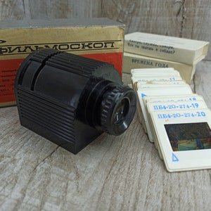 May include: A black plastic film projector with a lens and a slide tray. The projector is labeled "ФИЛЬМОСКОП" in Cyrillic lettering. The image also shows a stack of slides with the text "ПБ4-20-274-19" and "ПБ4-20-274-20" visible.
