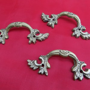 May include: Three ornate brass drawer pulls with a decorative leaf design. The pulls are in the shape of a half-circle with a raised center.