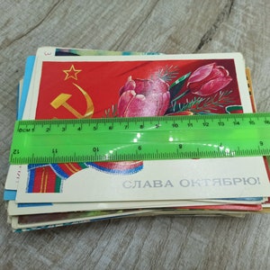 May include: A stack of vintage postcards, featuring Soviet-era imagery. The top card displays a red background with a gold hammer and sickle, a star, and a tulip. A green ruler shows the postcards are approximately 5 inches wide.