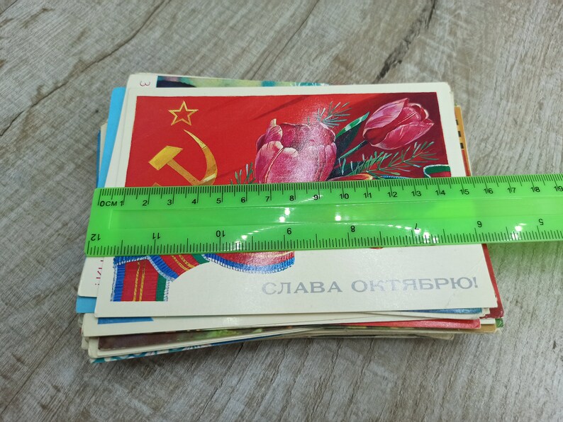 May include: A stack of vintage postcards, featuring Soviet-era imagery. The top card displays a red background with a gold hammer and sickle, a star, and a tulip. A green ruler shows the postcards are approximately 5 inches wide.