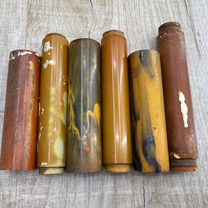 Può includere: Sei cilindri rotondi in legno di diversi colori. I cilindri sono tutti di lunghezza e diametro simili. Sono disposti in fila da sinistra a destra. I cilindri sono tutti di diverse tonalità di marrone e giallo.