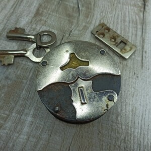May include: A vintage silver and black padlock with two keys. The padlock has a unique design with a keyhole in the center and a decorative pattern around the edges.