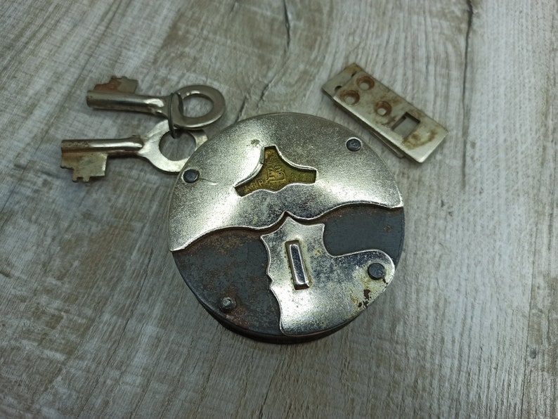 May include: A vintage silver and black padlock with two keys. The padlock has a unique design with a keyhole in the center and a decorative pattern around the edges.