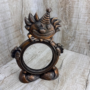 May include: A bronze-colored clown-shaped picture frame. The frame features a smiling clown face with a party hat atop its head. The clown's arms hold a circular frame, and the base has a diamond pattern.