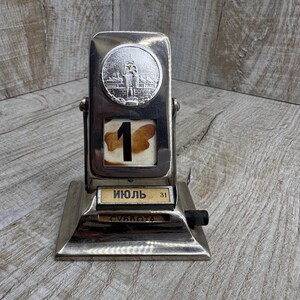 May include: A vintage silver-toned metal desk calendar displaying "July 1" in Russian. The calendar features a circular emblem and a black knob. The base is rectangular, and the background is a wooden surface.