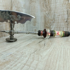 May include: A silver-toned, ornate butter knife with a decorative blade and a detailed handle. The handle transitions to a clear, cylindrical section filled with colorful floral elements. The knife rests on a light-colored wooden surface.