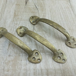 Op de afbeelding: Drie antieke messing kastdeurgrepen met een hartvormig ontwerp en vier decoratieve gaten. De grepen zijn in een rij op een licht houten oppervlak gerangschikt.