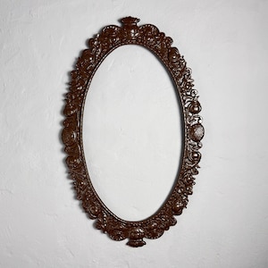 May include: An oval, ornate brown frame with intricate floral and scrollwork detailing. The frame is empty, with a white interior, and is mounted on a textured white wall. The frame has a decorative crown at the top and a similar design at the bottom.