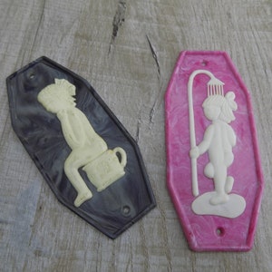 May include: Two vintage bathroom plaques. One is dark gray with a cream-colored figure seated on a chamber pot. The other is pink with a cream-colored figure under a shower head. Both have a hole for hanging.
