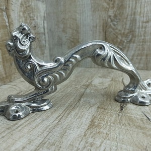 Pode incluir: Puxador de porta prateado e ornamentado com um design curvo. O puxador apresenta volutas decorativas e uma cabeça de animal estilizada e detalhada em uma extremidade. O puxador é feito de metal e tem uma estética vintage ou antiga.