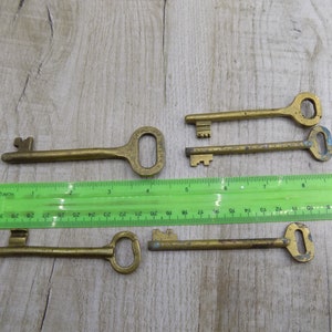 May include: Five antique brass keys of varying sizes and shapes, laid out on a wooden surface with a green ruler for scale.