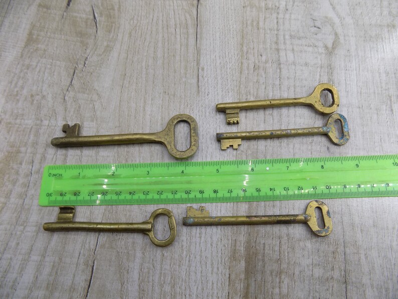 May include: Five antique brass keys of varying sizes and shapes, laid out on a wooden surface with a green ruler for scale.