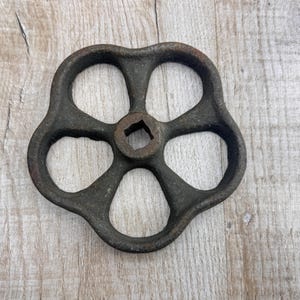 以下が含まれることがあります： 五つの丸みを帯びたスポークと中央の正方形の開口部を備えた、風化した濃い灰色の鋳鉄製バルブホイール。ホイールは明るい茶色の木製の表面に置かれ、その経年変化した質感と工業デザインを示しています。