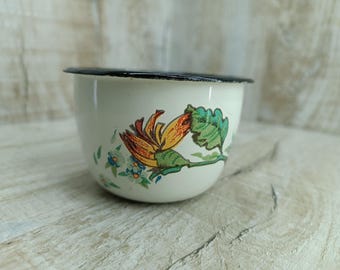May include: A white enamelware cup with a black rim and a floral design featuring yellow, orange, green, and blue flowers.