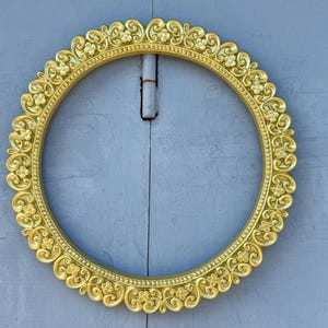 May include: A round, gold-colored picture frame with ornate floral and scroll detailing. The frame is set against a light blue background. The frame's design features repeating floral patterns and a scalloped edge.