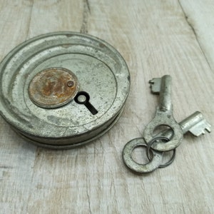 May include: A vintage metal lock with a keyhole and a round, silver key. The lock has a worn, aged appearance and is made of metal.