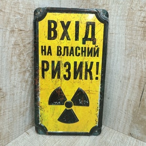 Può includere: Un cartello di avvertimento giallo e nero con il testo "ВХІД НА ВЛАСНИЙ РИЗИК!" che si traduce in "Entrata a proprio rischio!" in italiano. Il cartello presenta un simbolo di radiazione nero stilizzato.
