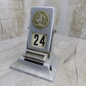 May include: A silver metal desk calendar with a flip-up design. The calendar features a round, embossed metal medallion with a figure sitting on a bench. The date '24' is displayed in a black font on a white background.
