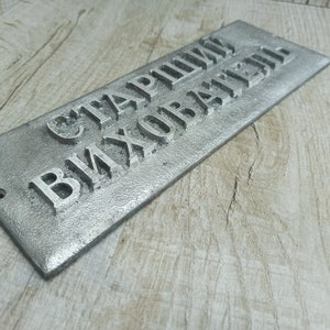 Puede incluir: Un letrero de metal plateado con el texto "Старший Вихователь" en letras en relieve.