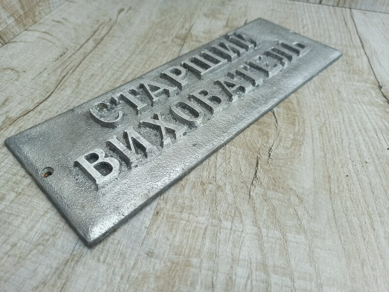 Puede incluir: Un letrero de metal plateado con el texto "Старший Вихователь" en letras en relieve.