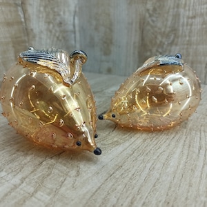 May include: Two glass hedgehog ornaments. The hedgehogs are a golden amber colour with small, raised bumps. They have black eyes and a silver and black leaf and mushroom detail on their backs. The ornaments are displayed on a light wood-grain surface.