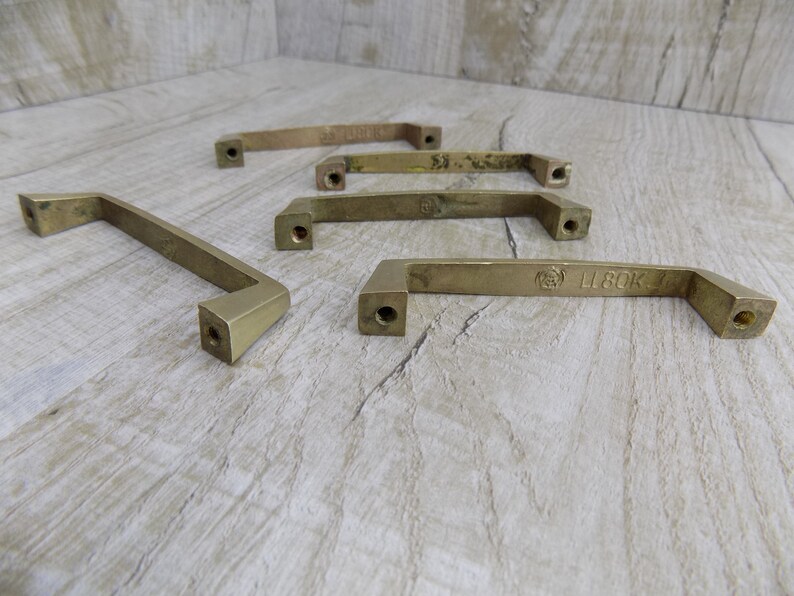 Op de afbeelding: Vijf messing kastgrepen met een rechthoekige vorm en afgeronde randen. De grepen hebben een geborstelde afwerking en zijn gemarkeerd met de tekst "L 80K 7".