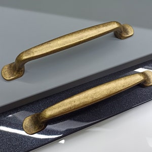 Peut inclure: Deux poignées de tiroir dorées, l'une sur l'autre. La poignée du haut est plus longue et a une forme plus arrondie. La poignée du bas est plus courte et a une forme plus rectangulaire.