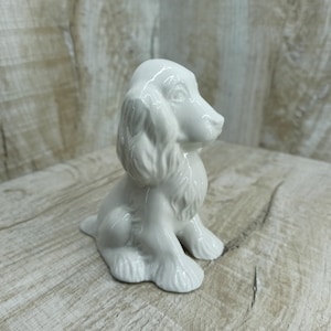 Puede incluir: Figurita de cerámica blanca de un perro cocker spaniel sentado. La figurita está detallada con pelo y tiene un aspecto realista.