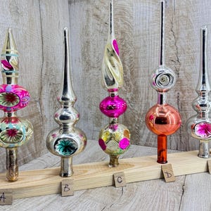 May include: Five glass Christmas tree toppers labelled A through E, arranged on a wooden stand. A features pink and teal accents, B is silver with a green centre, C has pink and gold patterns, D is orange and silver, and E is silver with pink.