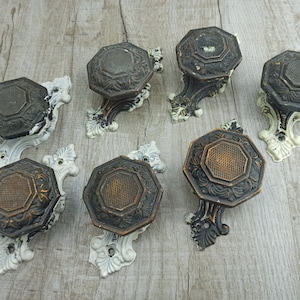 May include: Set of eight antique door knobs with ornate designs. The knobs are made of metal and have a dark brown finish. Each knob is attached to a white porcelain base.
