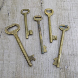 May include: Six antique brass keys of varying sizes and shapes, arranged on a light brown wooden surface.